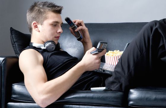 Un joven tumbado en un sofá utilizando un teléfono móvil, un ordenador portátil y un MP4