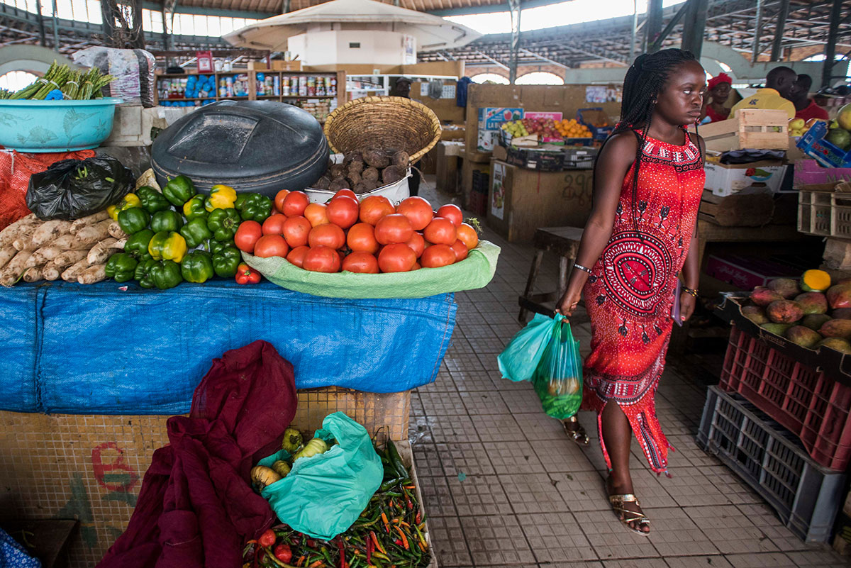 Una mujer pasa ante un puesto de verduras. A la izquierda, se puede ver mandioca, yuca o cassava, un cultivo más agradecido y resistente a la acidez del suelo y la sequía, del que también se puede fabricar harina
