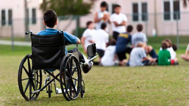 Un adolescente en silla de ruedas observa a sus compañeros jugando en el parque mientras le excluyen.