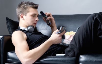 Un joven tumbado en un sof&aacute; utilizando un tel&eacute;fono m&oacute;vil, un ordenador port&aacute;til y un MP4. 