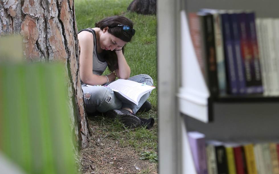 Una mujer lee un libro sentada en el césped, junto a las casetas de la Feria del Libro de Madrid, en el madrileño parque del Retiro.