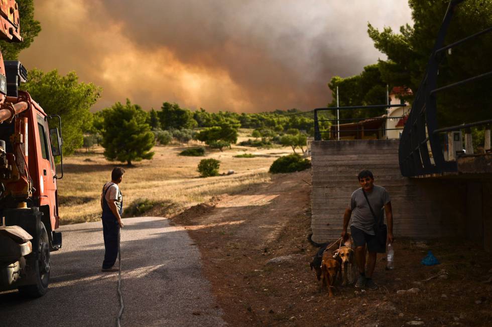 Un hombre sujeta una manguera y otro camina con sus perros cerca de un incendio en Grecia.