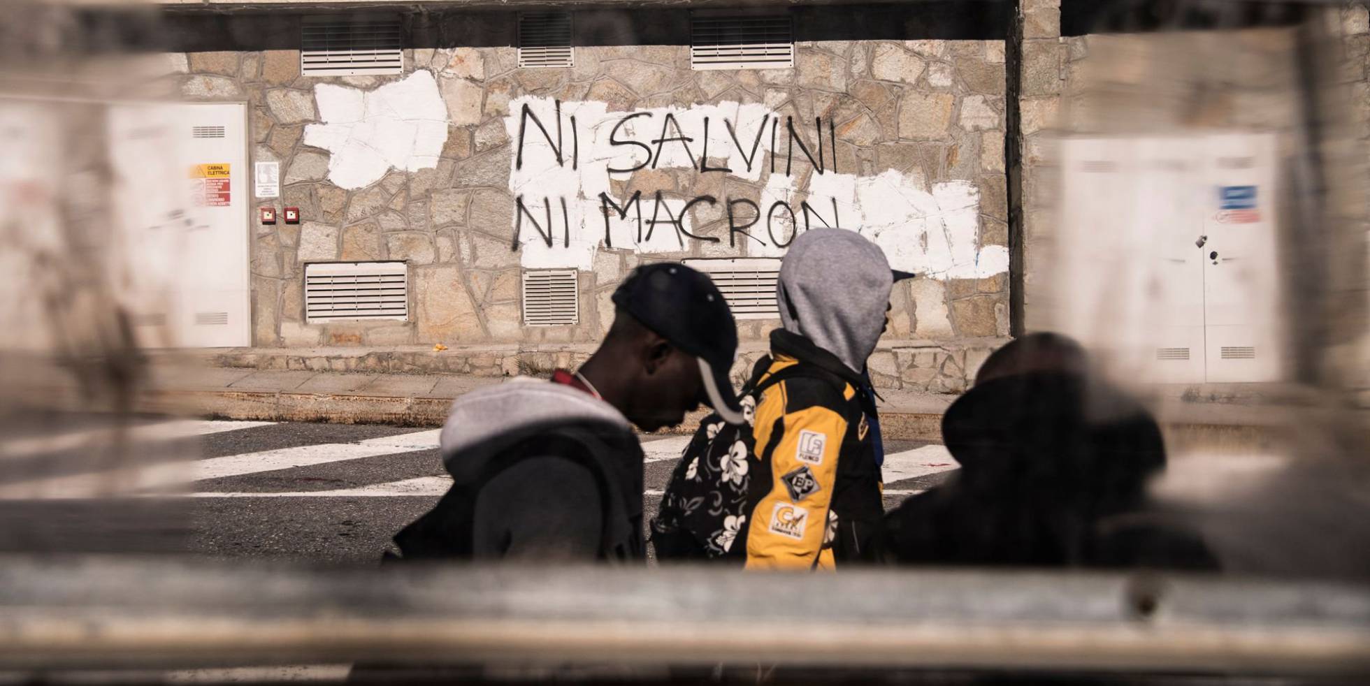 Tres migrantes pasa junto a un grafiti en el que puede leerse 'Ni Salvini, ni Macron, en la localidad de Claviere (italia).