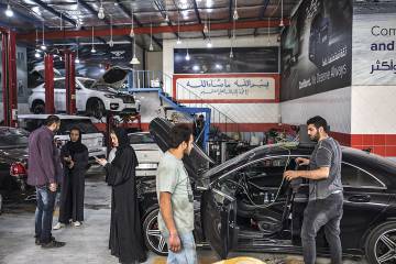 Elham, dentro de seu veículo, observa Saja dar instruções a Yasin, e pergunta ao mecânico quando poderá passar para pegar o carro.