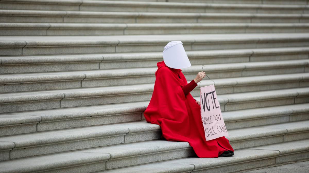La activista Kelli Midgley disfrazada de El cuento de la criada, sostiene una pancarta en la que se lee