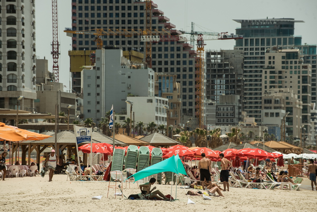 Una playa de Tel Aviv con sombrillas y tumbonas de alquiler municipales junto al paseo marítimo, al fondo hoteles y apartamentos de lujo